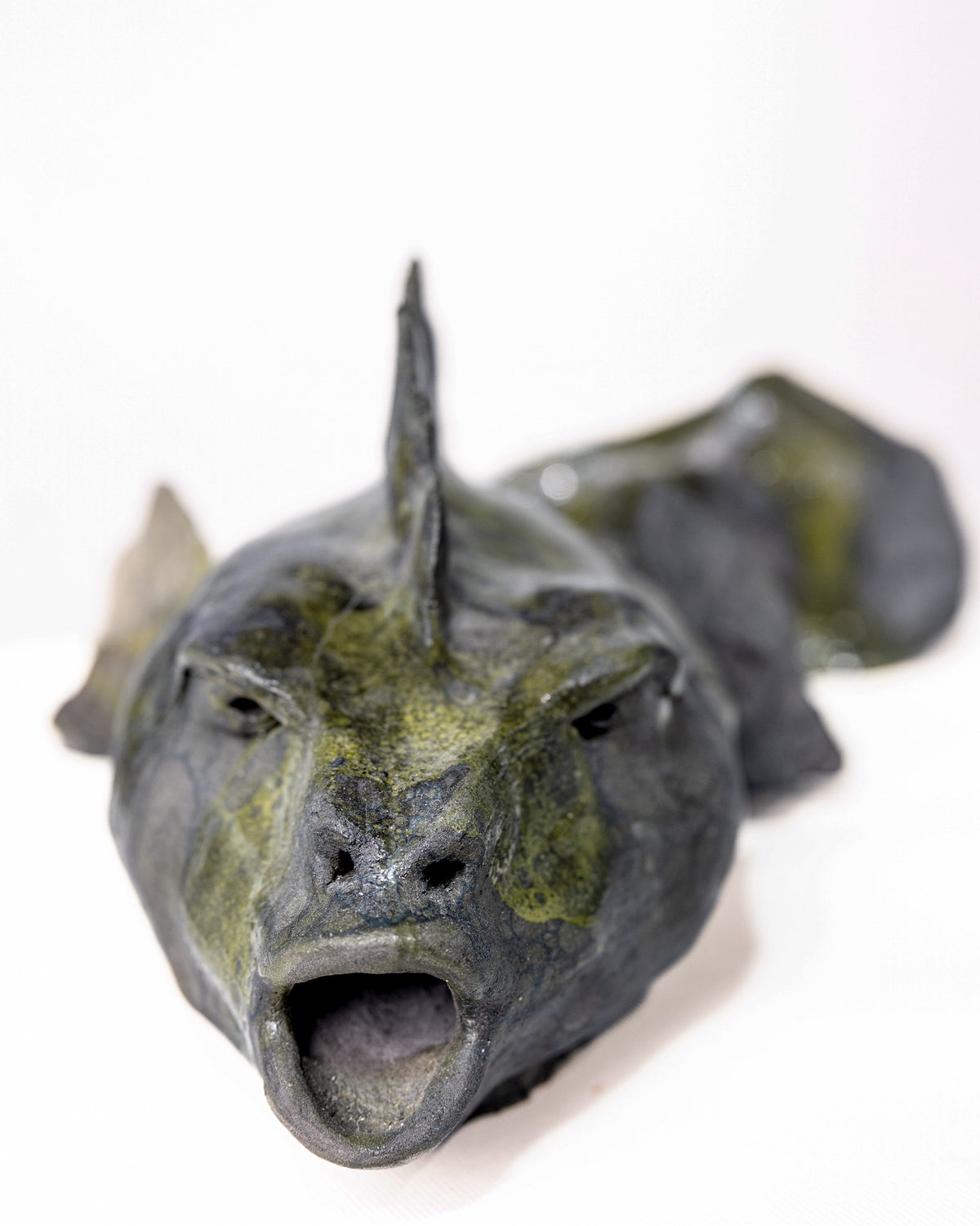 A handmade ceramic sculpture of a low, elongated fish resting on a flat surface, featuring an open mouth, a jagged dorsal fin along its back, and a dark slate grey glaze with pooling olive green patches, set against a plain white background.