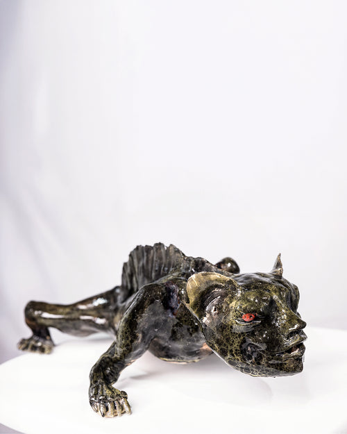A handcrafted ceramic sculpture of a low-crouching creature with a broad, flattened face and a finned spine, crawling forward with a slick, volcanic glaze in deep olive, black, and mossy green tones, featuring glowing red eyes beneath a heavy brow, set against a plain white background.