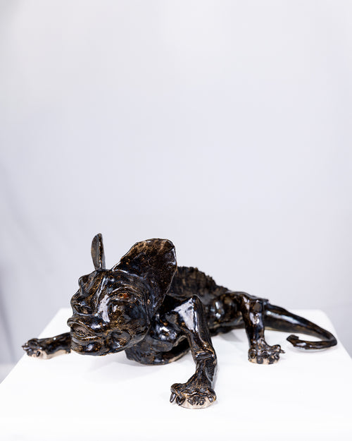 A handcrafted dark ceramic sculpture of a low, crouching creature stretching forward on a white surface, featuring an elongated human-like face, oversized ears, a ridged spine, and a curling tail, with a glossy glaze reflecting light in deep tones.