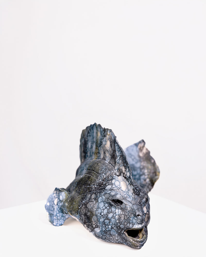 handcrafted ceramic sculpture of a wide-bodied fish with a broad head and open mouth, featuring a dominant fin and a textured, cellular bubble glaze in dark grey, black, and blue tones against a plain white background.