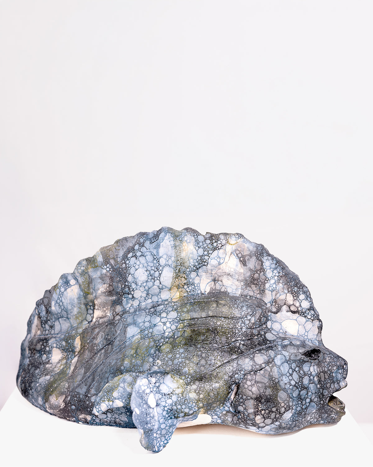 A handcrafted ceramic sculpture of a wide-bodied fish with a broad head and open mouth, featuring a dominant fin and a textured, cellular bubble glaze in dark grey, black, and blue tones against a plain white background.