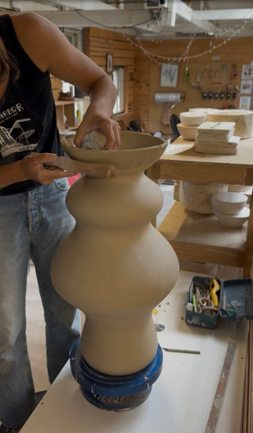 A person in a pottery studio shaping a large, stacked bulbous ceramic vase on a banding wheel, working on the flared rim with a tool against a rustic wooden interior backdrop.