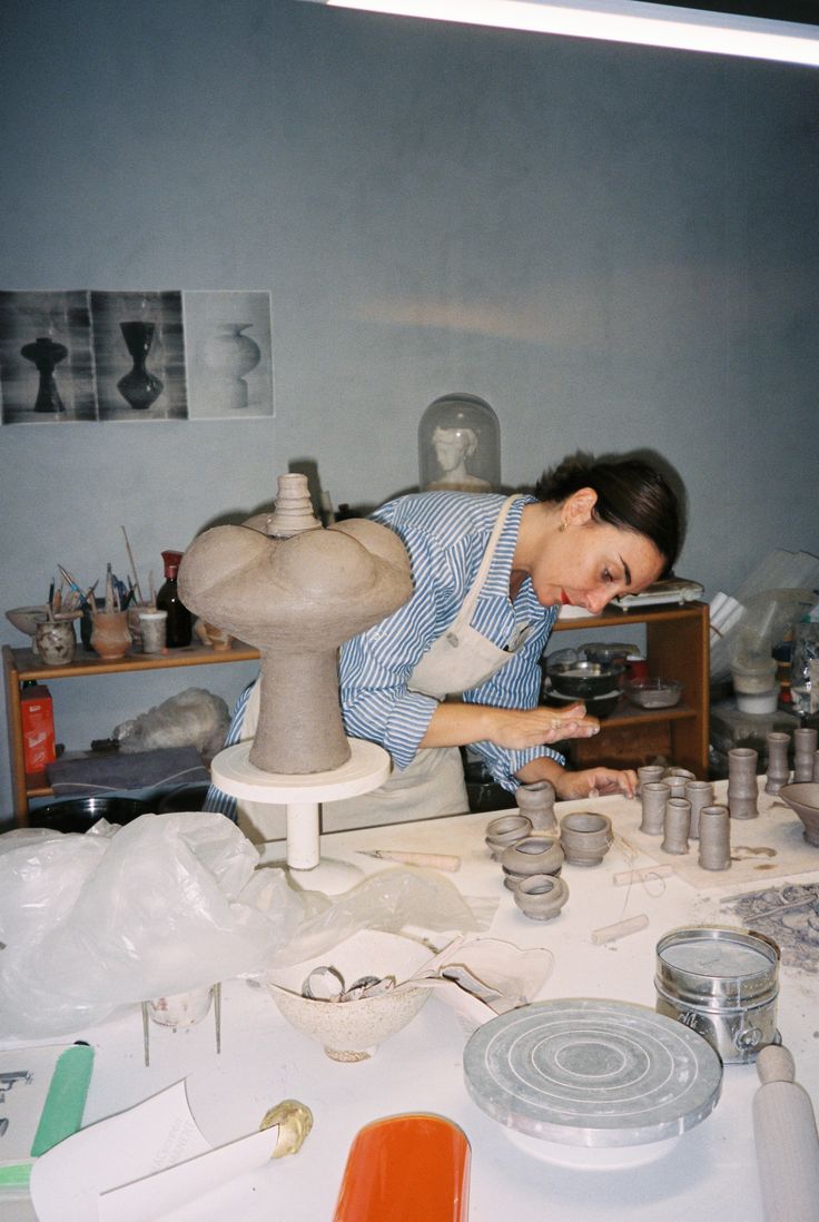 A person in a pottery studio shaping a large, stacked bulbous ceramic vase on a banding wheel, working on the flared rim with a tool against a rustic wooden interior backdrop.