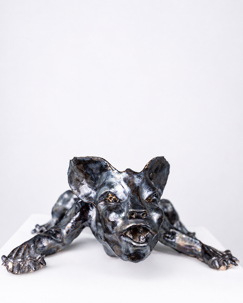 A handcrafted ceramic sculpture of a low, crouching creature with wide ears and an aggressive, open-mouthed expression, glazed in a lustrous blend of black and silver tones that shift between shadow and metallic sheen, set against a plain white background.