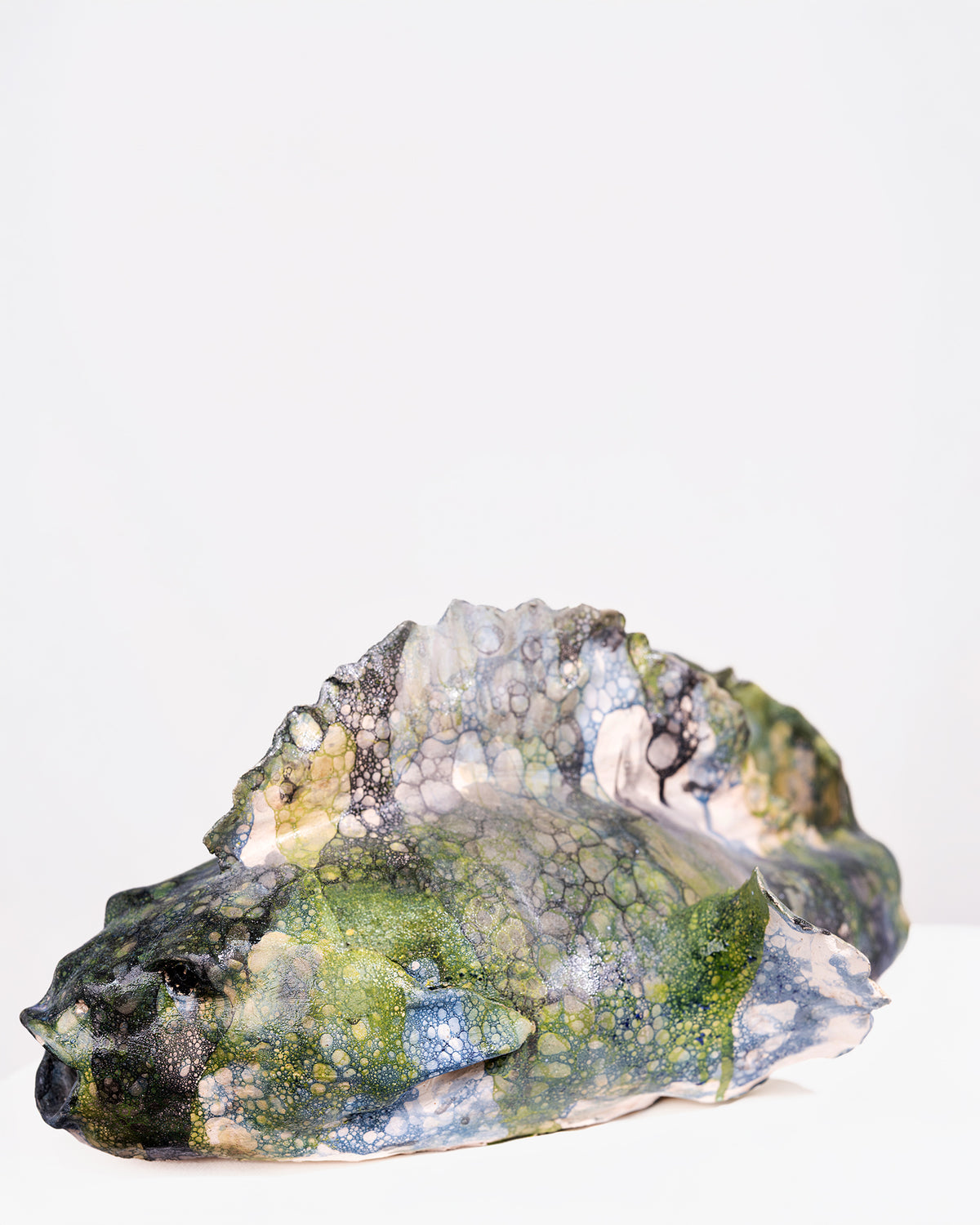 A handmade ceramic sculpture of a large fish with a wide, rounded body and an open mouth, glazed in a distinctive bubbled cellular pattern of olive green, yellow-green, pale blue, and dark grey, set against a plain white background.