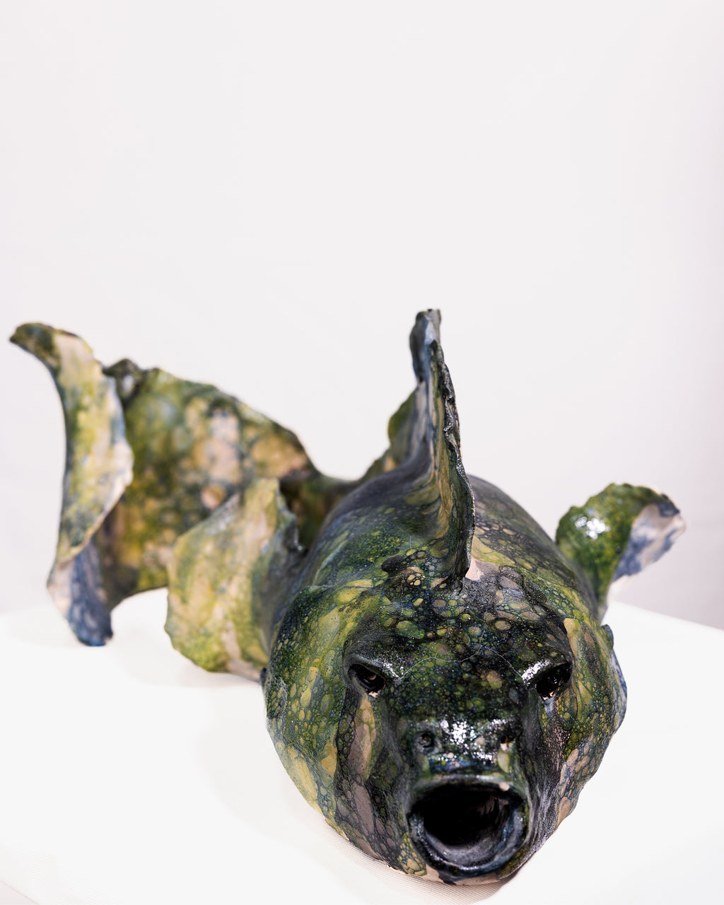 A handmade ceramic sculpture of a large fish with a wide, rounded body and an open mouth, glazed in a distinctive bubbled cellular pattern of olive green, yellow-green, pale blue, and dark grey, set against a plain white background.