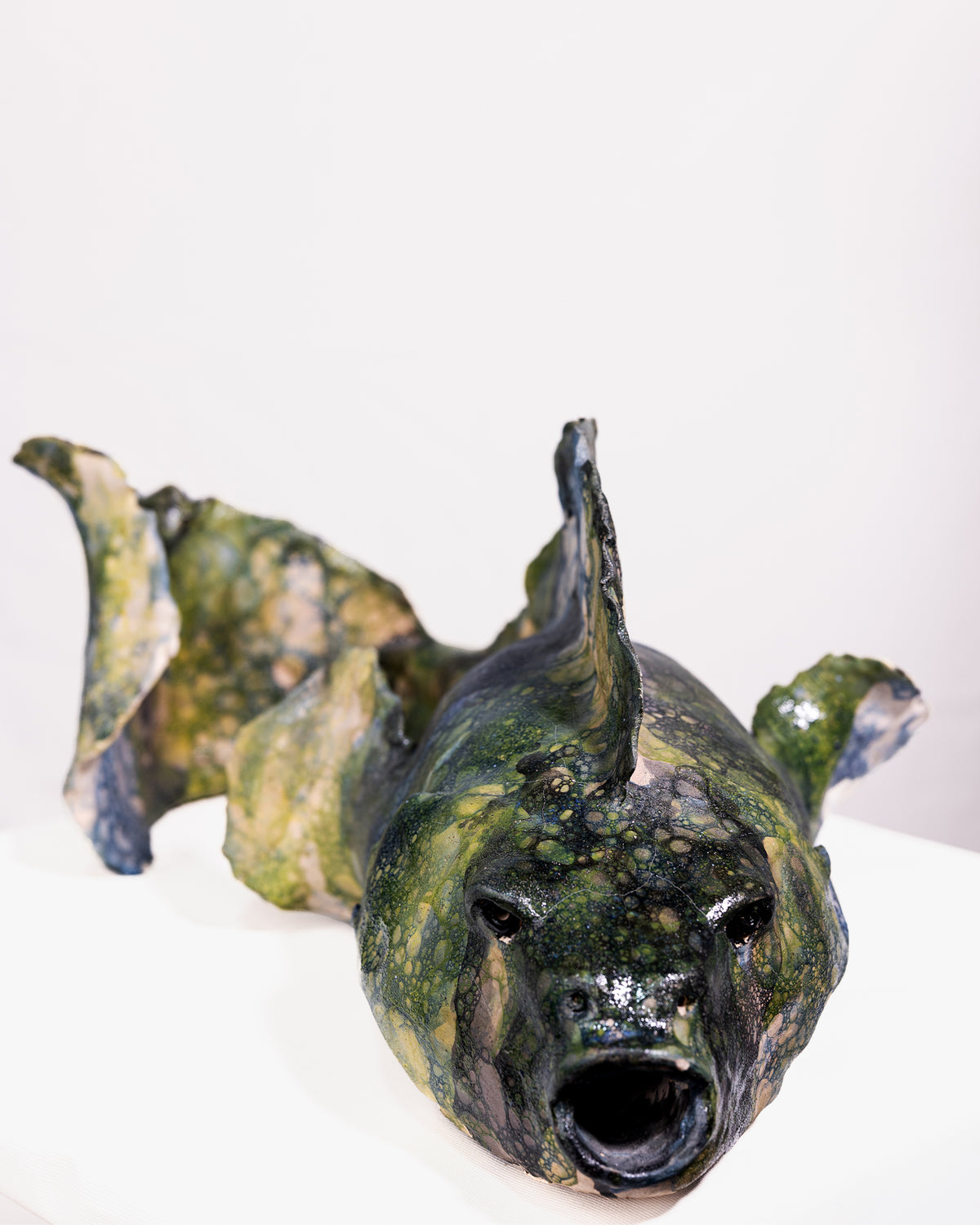 A handmade ceramic sculpture of a large fish with a wide, rounded body and an open mouth, glazed in a distinctive bubbled cellular pattern of olive green, yellow-green, pale blue, and dark grey, set against a plain white background.