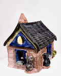 A glossy, house-shaped ceramic object with a dark blue and brown glaze, featuring textured brick details at the base, a pitched roof with a tall chimney, and small accent vases by the entrance.