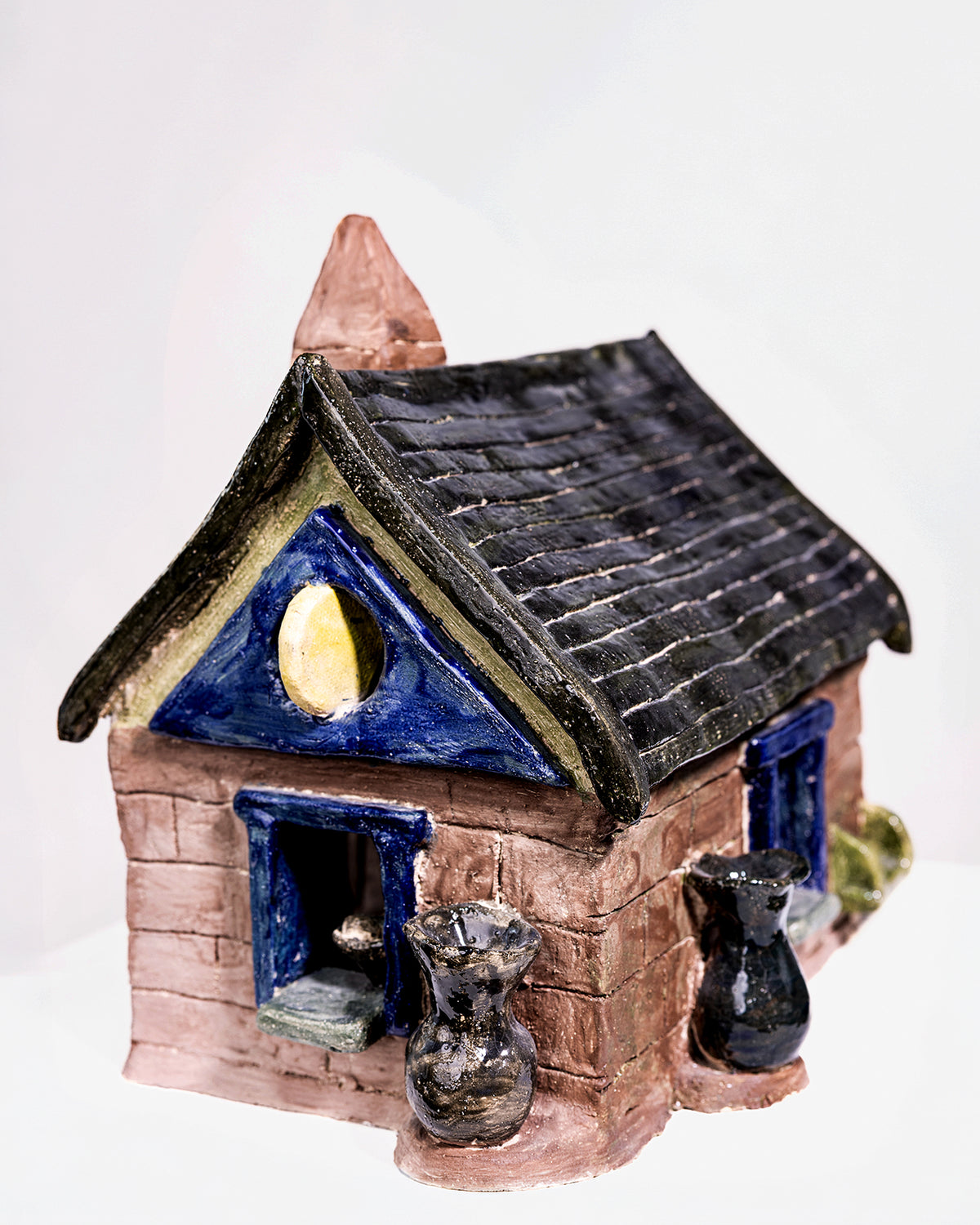 A glossy, house-shaped ceramic object with a dark blue and brown glaze, featuring textured brick details at the base, a pitched roof with a tall chimney, and small accent vases by the entrance.