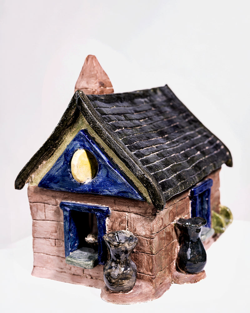 A glossy, house-shaped ceramic object with a dark blue and brown glaze, featuring textured brick details at the base, a pitched roof with a tall chimney, and small accent vases by the entrance.