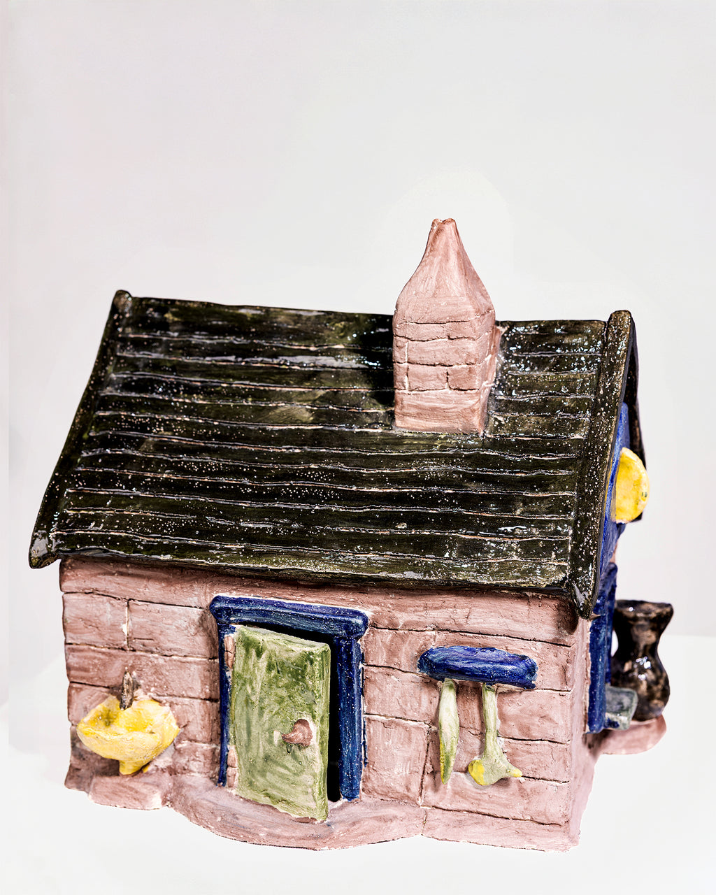 A handmade ceramic cottage figurine with a pinkish-beige brick-textured body, a dark gray shingled roof, a pink chimney, a green door, and blue trim details, shown against a plain white background.