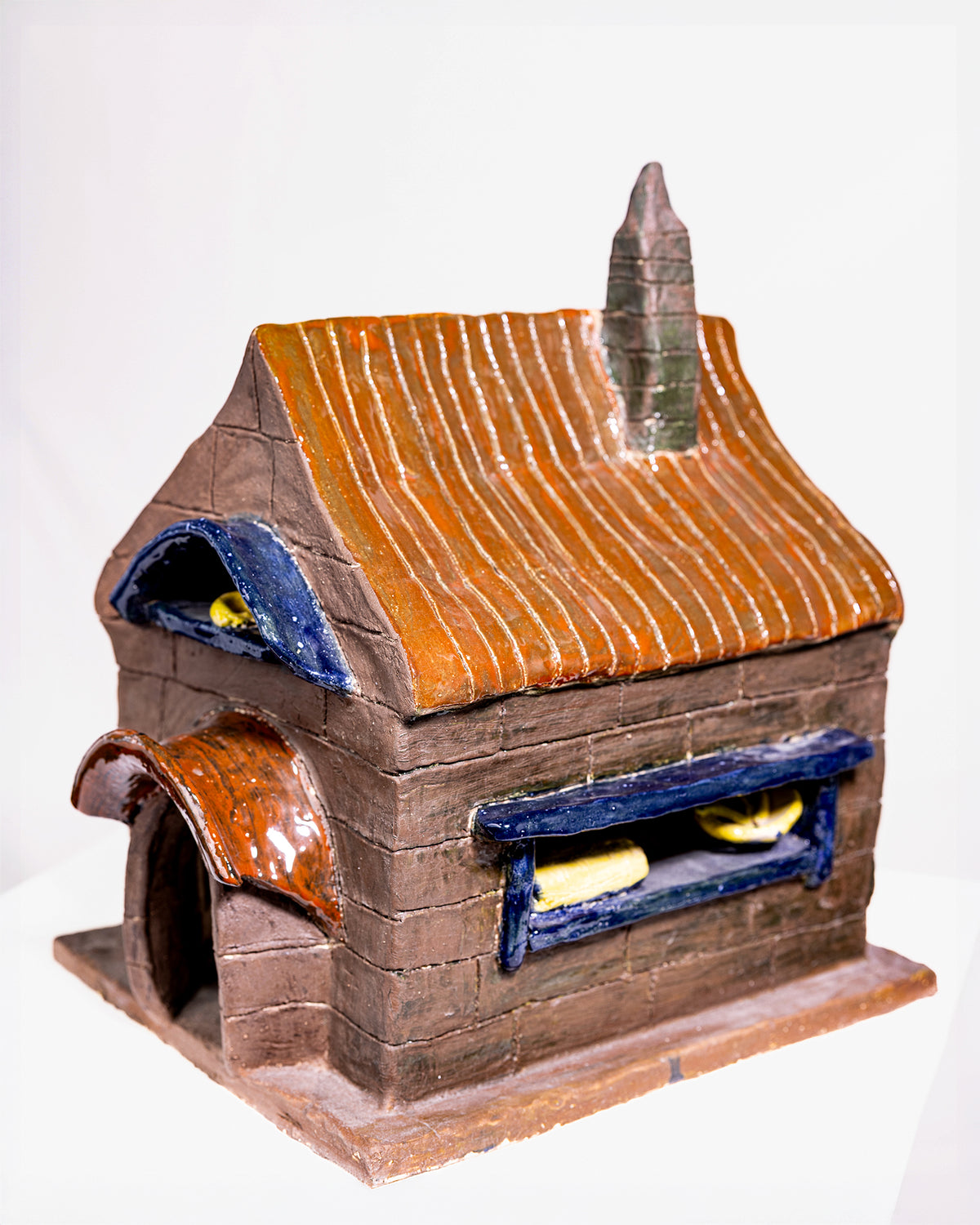 A handmade ceramic miniature house or oven structure with a brown brick-textured body, orange-brown striped roof, blue window trim, and a small chimney, photographed on a plain white background.