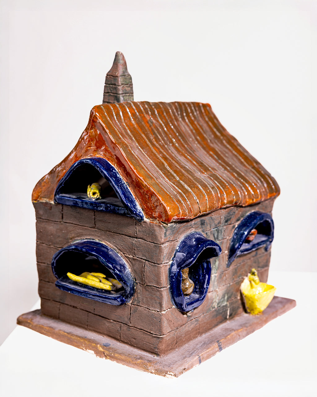 A handcrafted ceramic birdhouse shaped like a small house with a brown textured brick body, a brown-orange striped roof, and blue glazed trim around the windows and chimney.