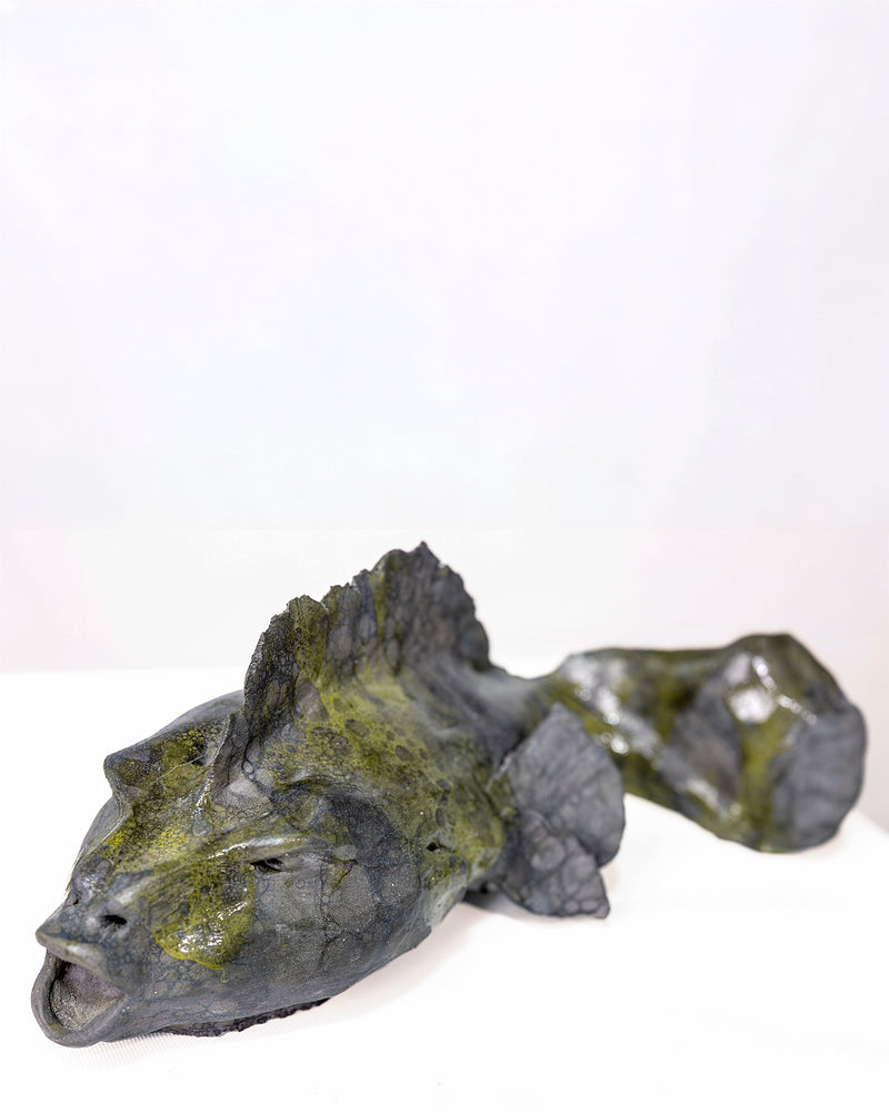 A handmade ceramic sculpture of a low, elongated fish resting on a flat surface, featuring an open mouth, a jagged dorsal fin along its back, and a dark slate grey glaze with pooling olive green patches, set against a plain white background.