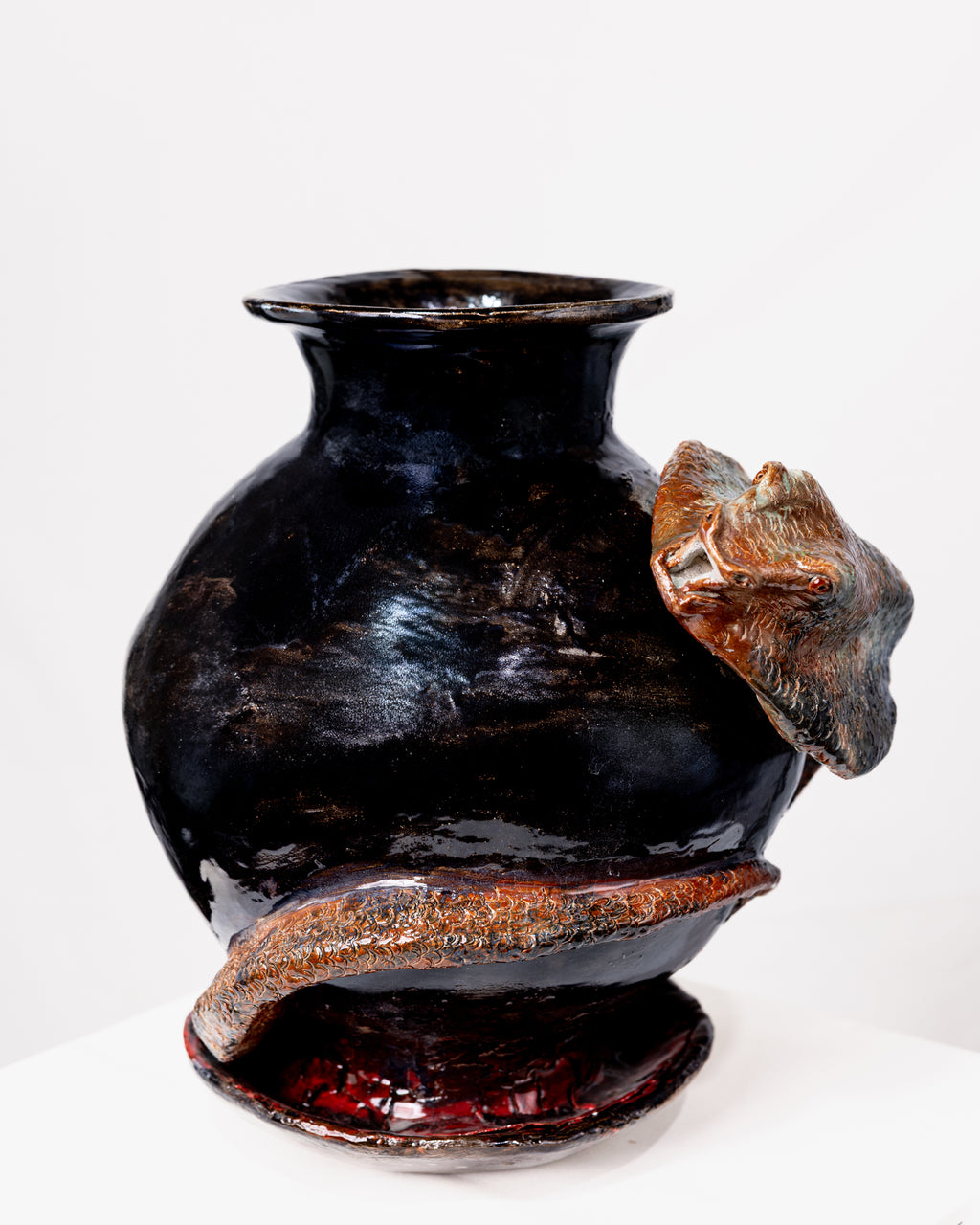 A round, glossy black ceramic vase with a flared rim, wrapped by a textured snake with scaled earthy orange-brown and blue-green details, set against a white background.