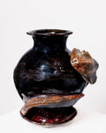 A round, glossy black ceramic vase with a flared rim, wrapped by a textured snake with scaled earthy orange-brown and blue-green details, set against a white background.