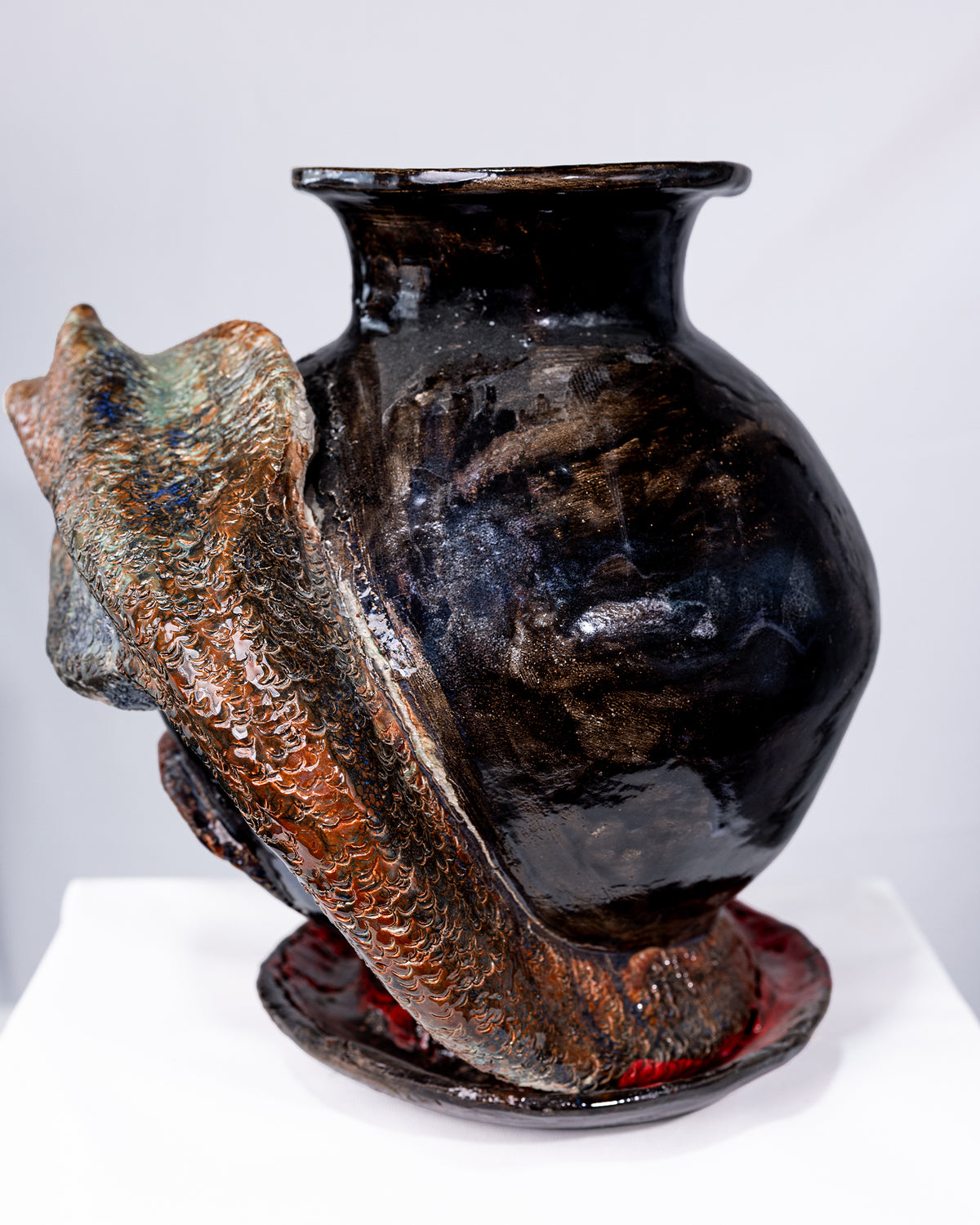 A round, glossy black ceramic vase with a flared rim, wrapped by a textured snake with scaled earthy orange-brown and blue-green details, set against a white background.