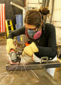 A woman wearing safety glasses, a pink respirator mask, and protective gloves, using an angle grinder on a metal bar on a workbench, creating a bright spray of sparks in a workshop setting.