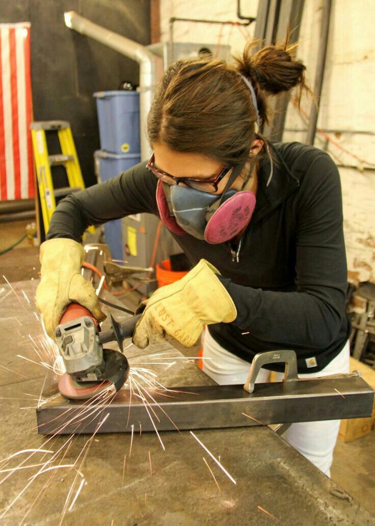 A woman wearing safety glasses, a pink respirator mask, and protective gloves, using an angle grinder on a metal bar on a workbench, creating a bright spray of sparks in a workshop setting.