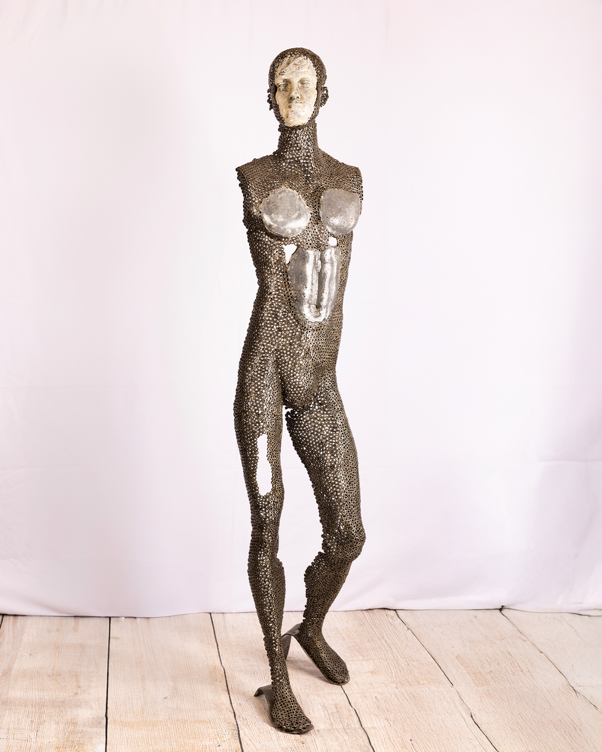 A life-sized figurative sculpture of a standing human figure, with its body constructed from dark iron nuts and mesh, contrasting with smooth, pale lead emerging across the face, chest, and torso, standing on a wooden floor against a white backdrop.