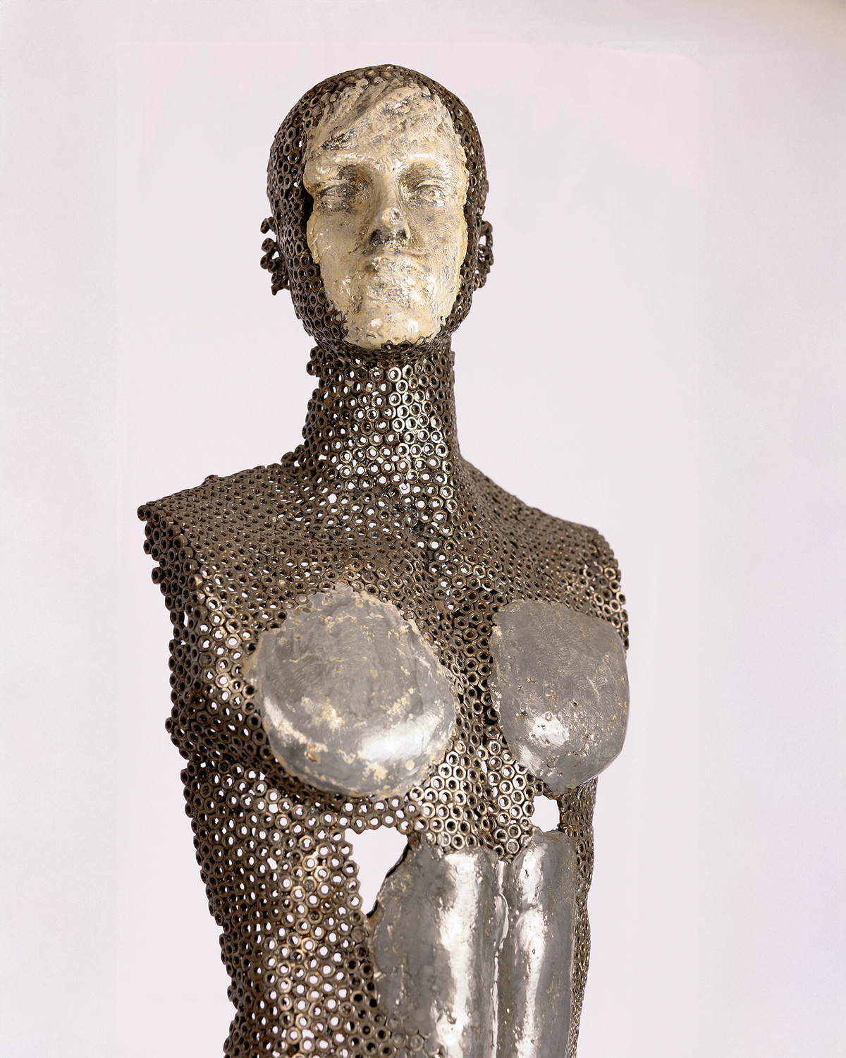 A life-sized figurative sculpture of a standing human figure, with its body constructed from dark iron nuts and mesh, contrasting with smooth, pale lead emerging across the face, chest, and torso, standing on a wooden floor against a white backdrop.