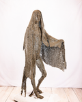 A life-sized figurative sculpture of a veiled figure moving forward, constructed entirely from welded iron nuts and mesh cascading from head to ground, with one arm reaching beneath the veil, set against a plain background.