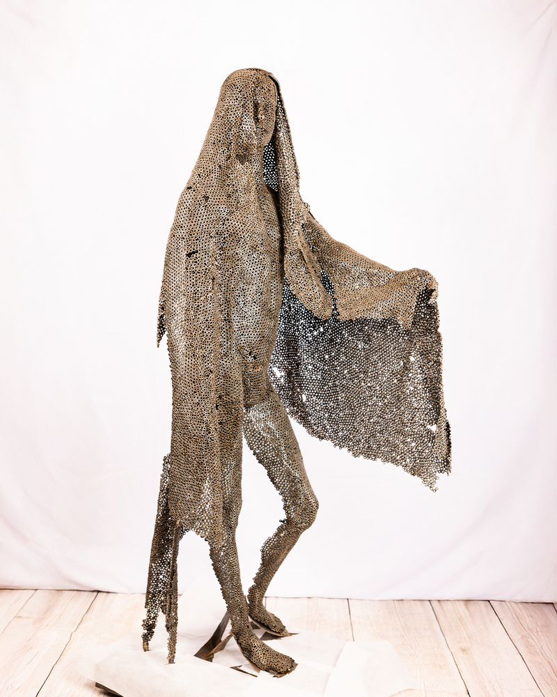 A life-sized figurative sculpture of a veiled figure moving forward, constructed entirely from welded iron nuts and mesh cascading from head to ground, with one arm reaching beneath the veil, set against a plain background.