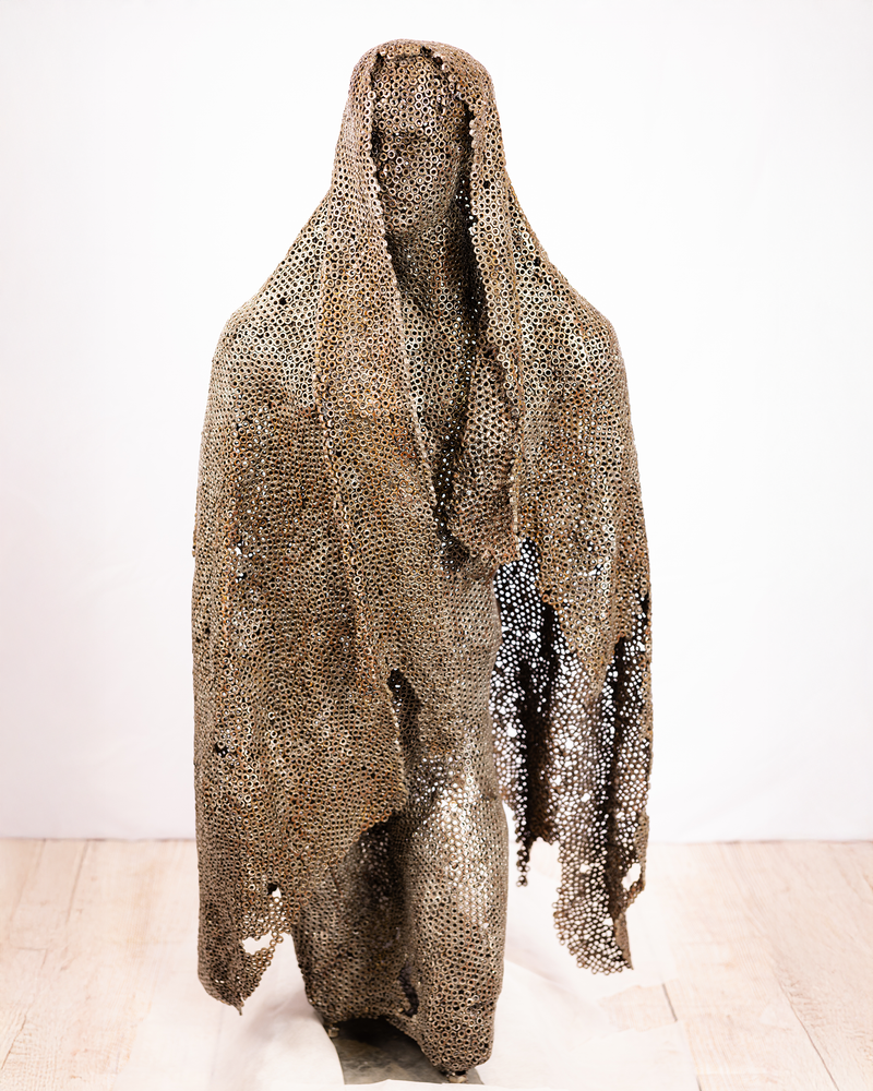 A sculpture of a kneeling figure enveloped in a sweeping cloak constructed entirely from welded dark iron nuts, with the face absorbed into the textured mesh lattice, set against a plain background.