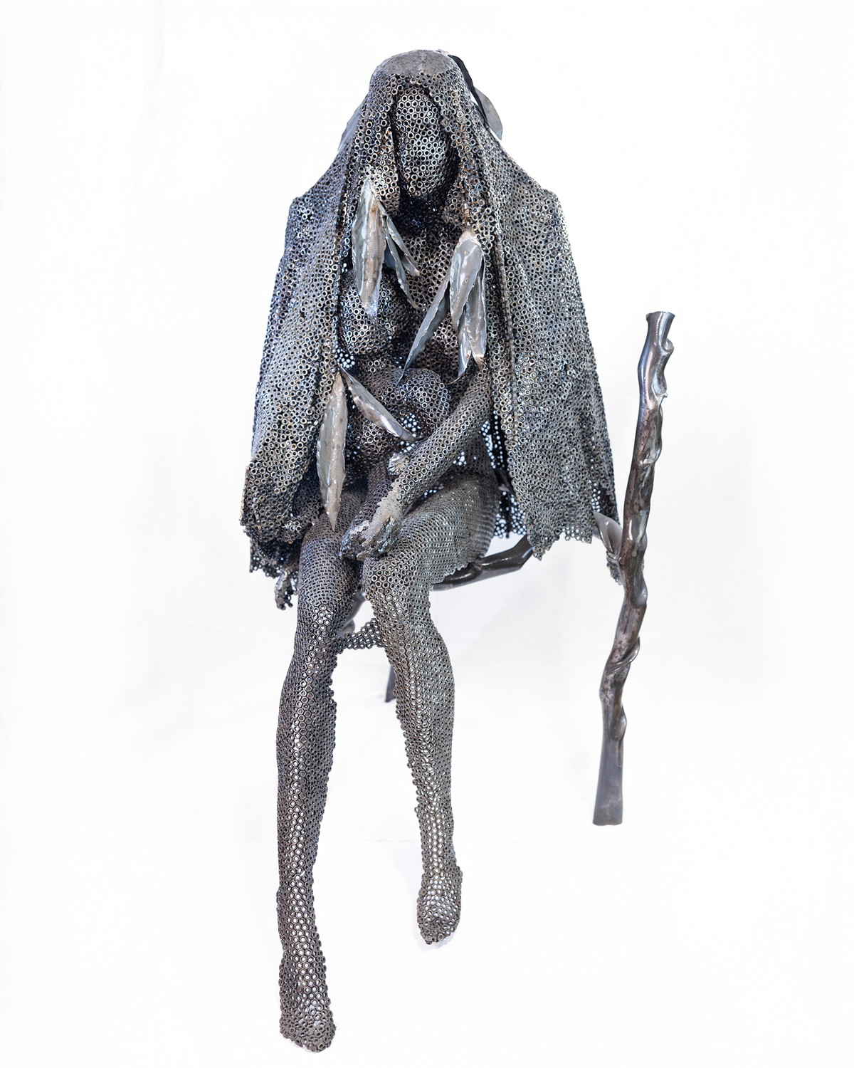 A life-sized sculpture of a seated mother holding her son close, constructed entirely from a lattice of welded dark iron nuts with a walking stick at her side, set against a plain background.