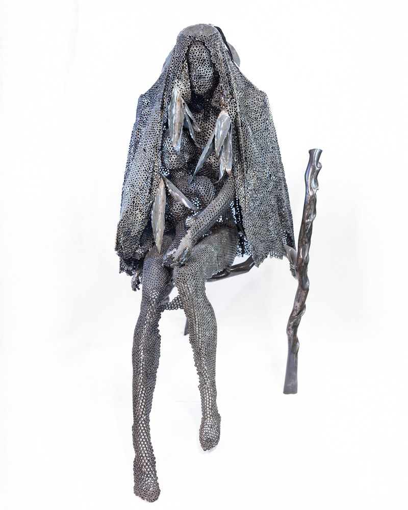 A life-sized sculpture of a seated mother holding her son close, constructed entirely from a lattice of welded dark iron nuts with a walking stick at her side, set against a plain background.