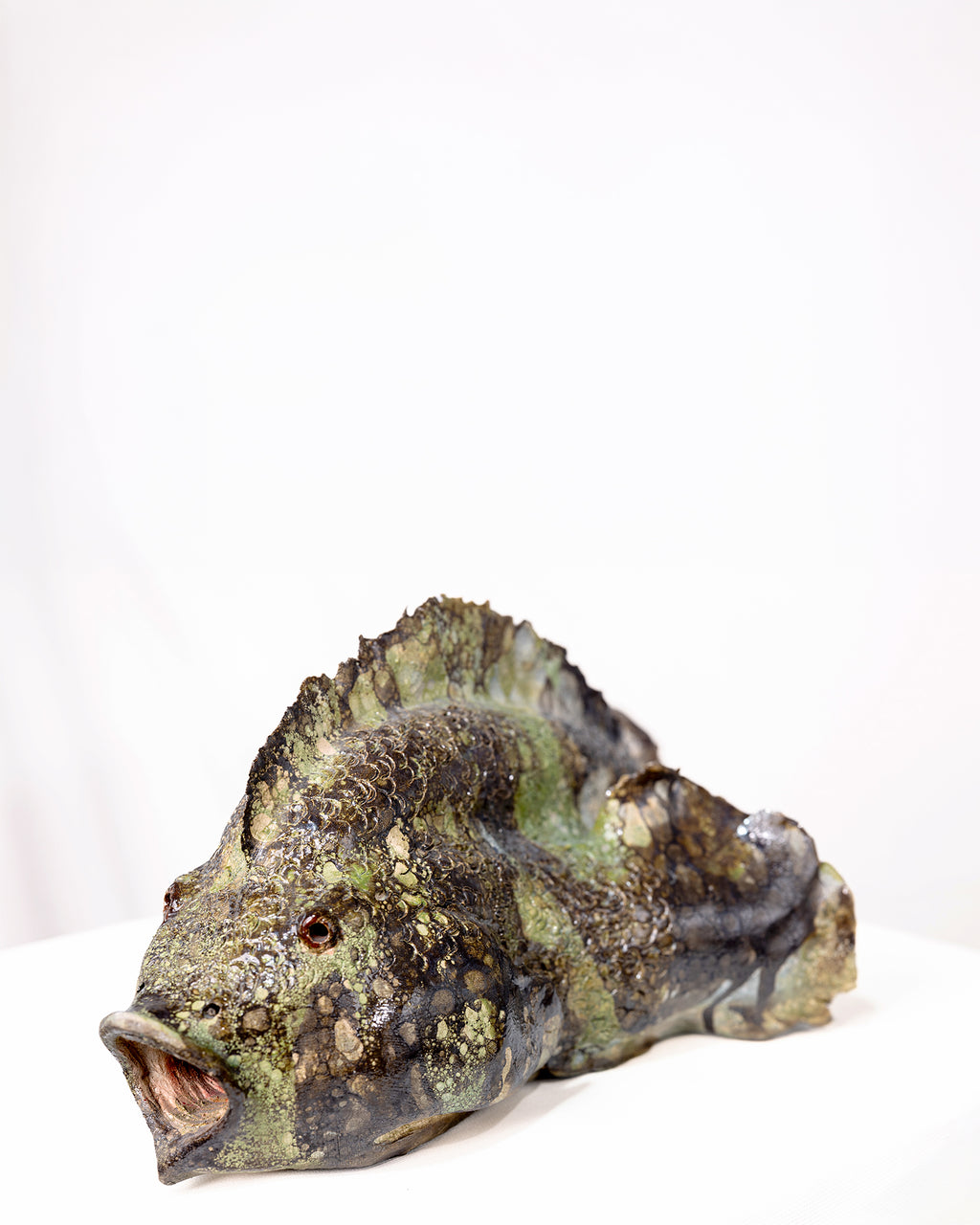 A handmade ceramic sculpture of a large fish with an open mouth and a densely textured body, glazed in dark brown, olive green, and cream tones against a plain white background.