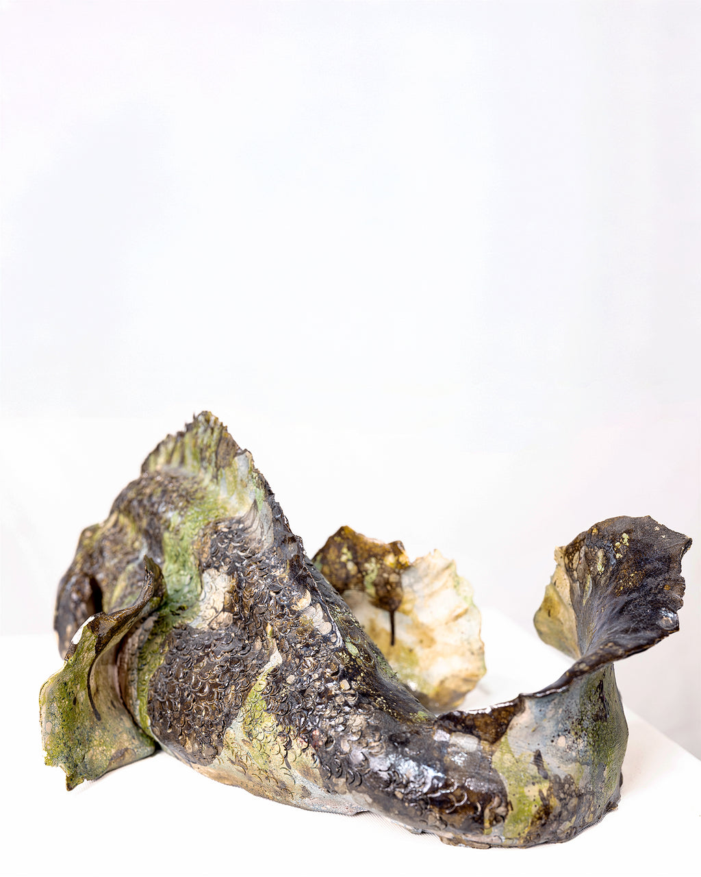 A sculptural fish-shaped object with textured, mossy surfaces in dark charcoal, olive green, and cream, photographed on a plain white background.