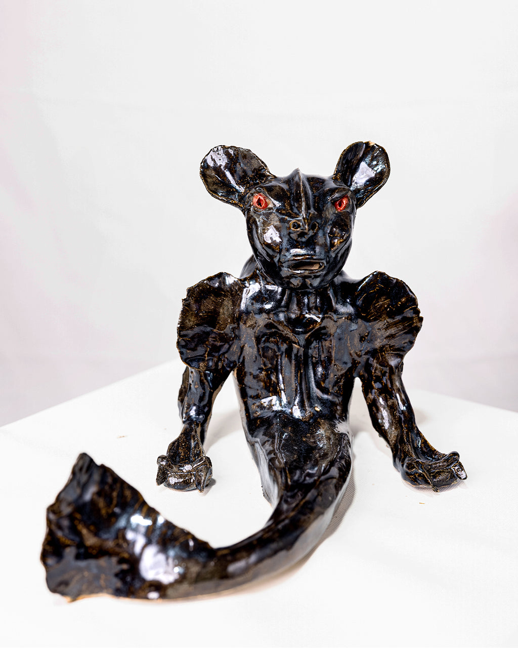 A handmade ceramic sculpture of a hybrid fantasy creature sitting upright with clawed arms, featuring a glossy deep black glaze with subtle gold undertones and striking red eyes, tapering into a long fish tail with a splayed fin against a white background.