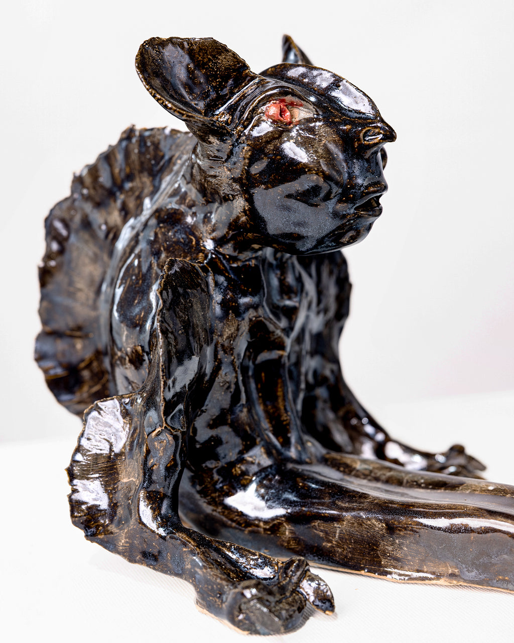 A handmade ceramic sculpture of a hybrid fantasy creature sitting upright with clawed arms, featuring a glossy deep black glaze with subtle gold undertones and striking red eyes, tapering into a long fish tail with a splayed fin against a white background.