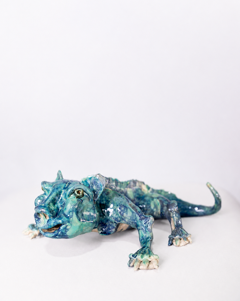 A handcrafted ceramic sculpture of a low-crouching creature, part lizard and part predator, with splayed claws and a slightly parted jaw, glazed in a shifting wash of deep teal and ocean blue tones against a plain background.
