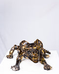 A handmade ceramic sculpture of a low, crouching creature with splayed arms and an intense expression, glazed in a striking pattern of deep black and gold pooling gloss, set against a plain white background.