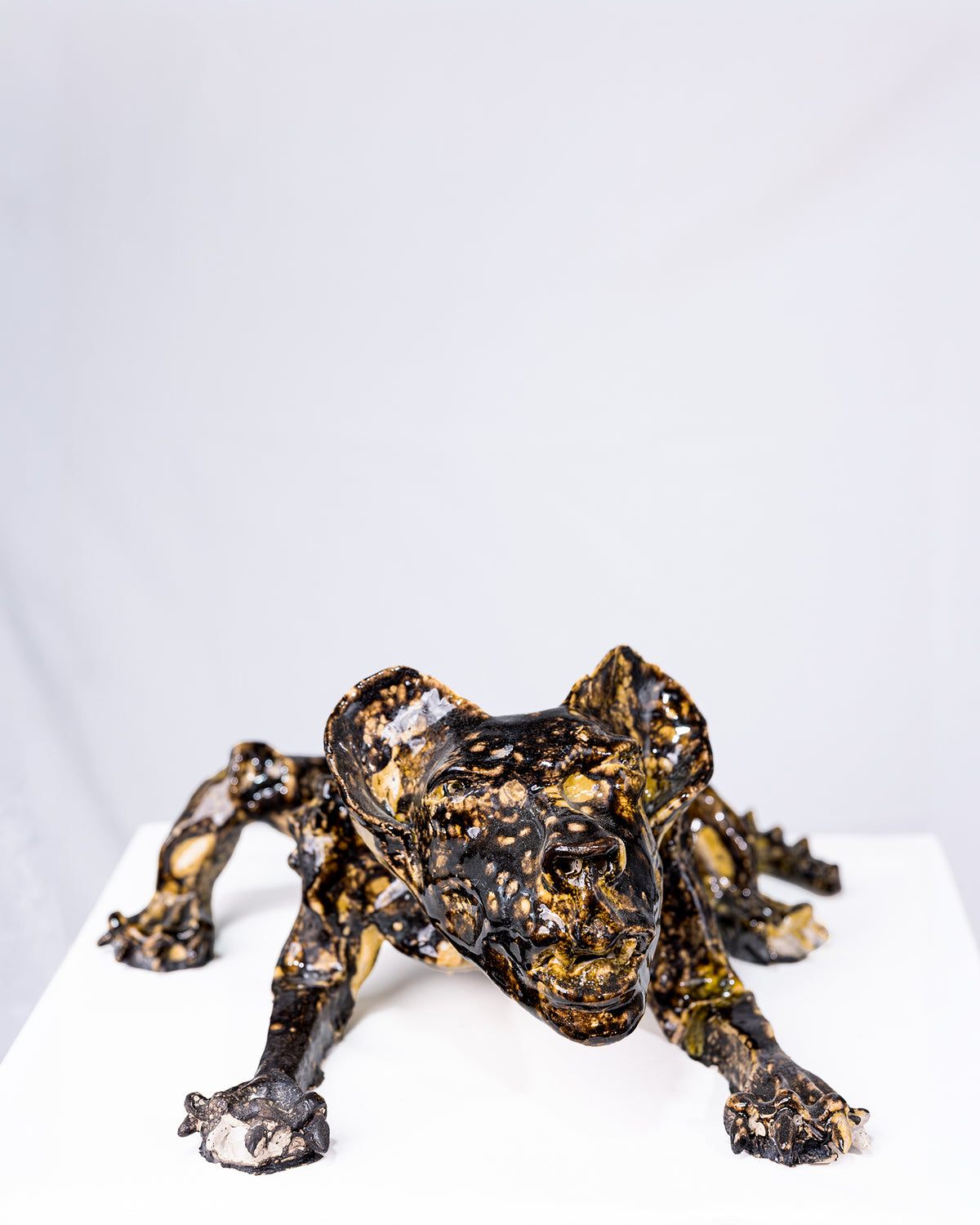 A handmade ceramic sculpture of a low, crouching creature with splayed arms and an intense expression, glazed in a striking pattern of deep black and gold pooling gloss, set against a plain white background.