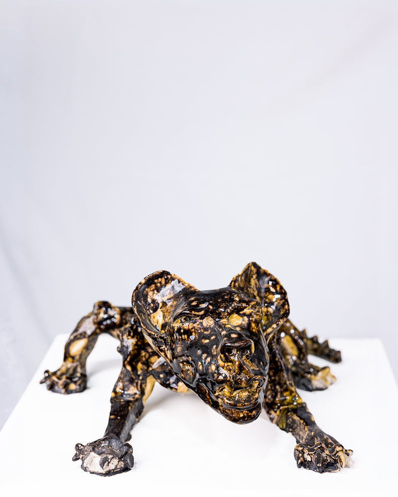 A handmade ceramic sculpture of a low, crouching creature with splayed arms and an intense expression, glazed in a striking pattern of deep black and gold pooling gloss, set against a plain white background.