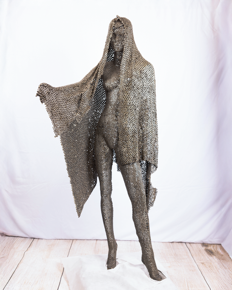 A life-sized figurative sculpture of a tall, standing female figure draped in a wide, billowing veil, constructed entirely from a continuous lattice of welded dark iron nuts against a neutral background.