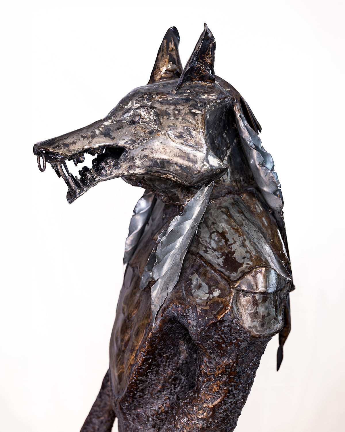 A textured metal sculpture of a wolf's head and upper body in profile, featuring layered metallic plates with a dark, weathered finish and polished silver highlights, set against a plain white background.