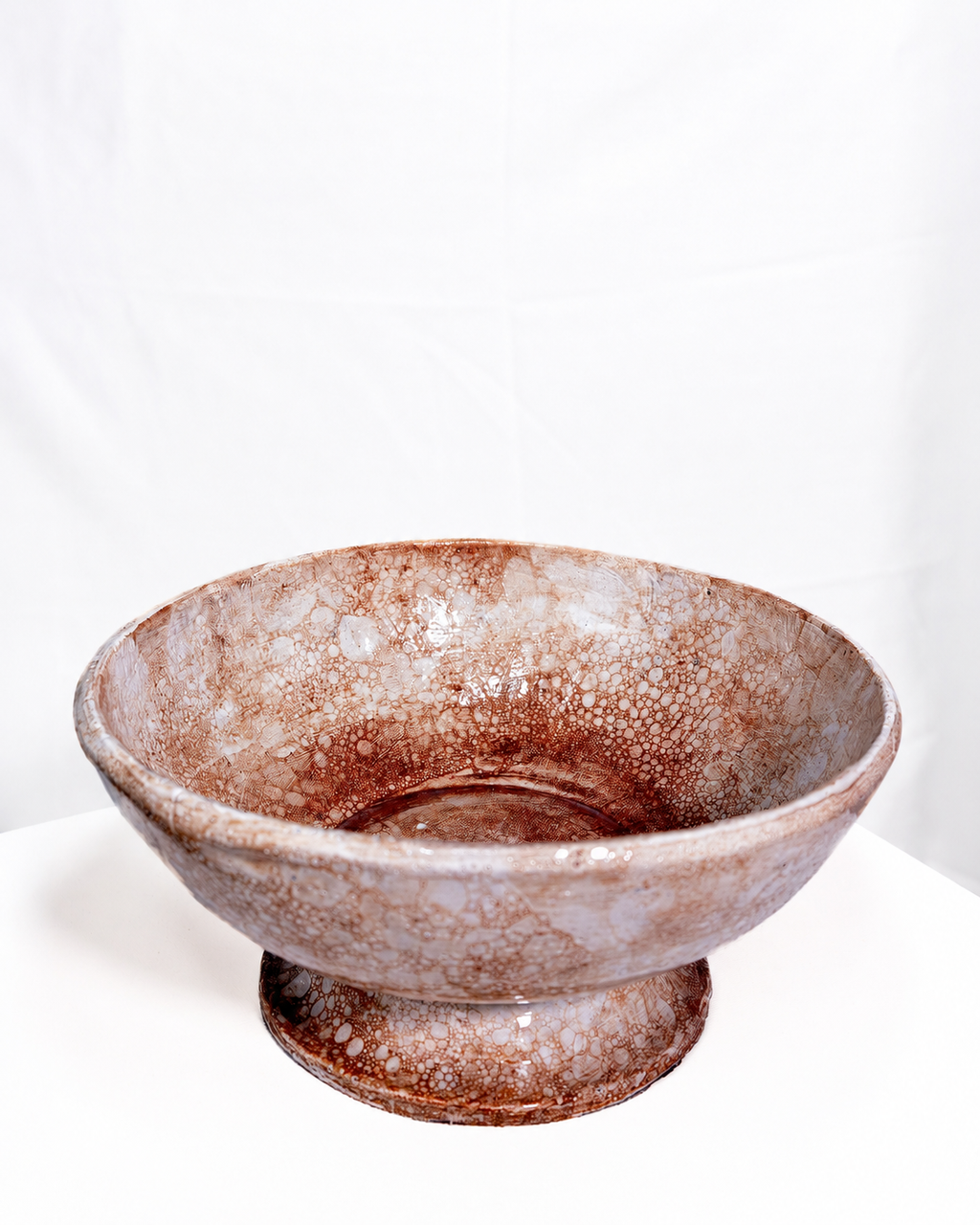 A handmade ceramic pedestal bowl with a textured, mottled glaze in earthy brown and beige tones, set against a plain white backdrop.