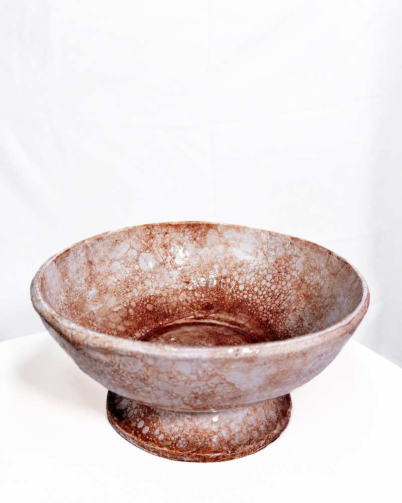 A handmade ceramic pedestal bowl with a textured, mottled glaze in earthy brown and beige tones, set against a plain white backdrop.