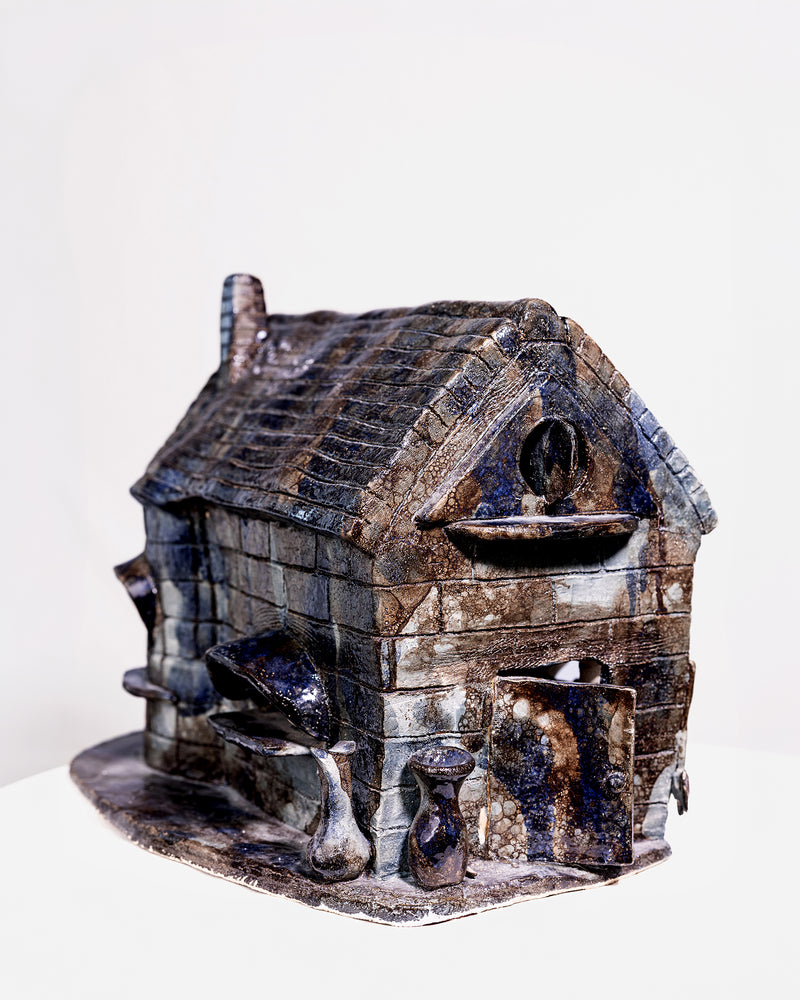 A glossy, house-shaped ceramic object with a dark blue and brown glaze. It features detailed brick-like texturing at the base, a tiled roof with a tall chimney, and a large curved barrel structure on the side, isolated against a white studio background.