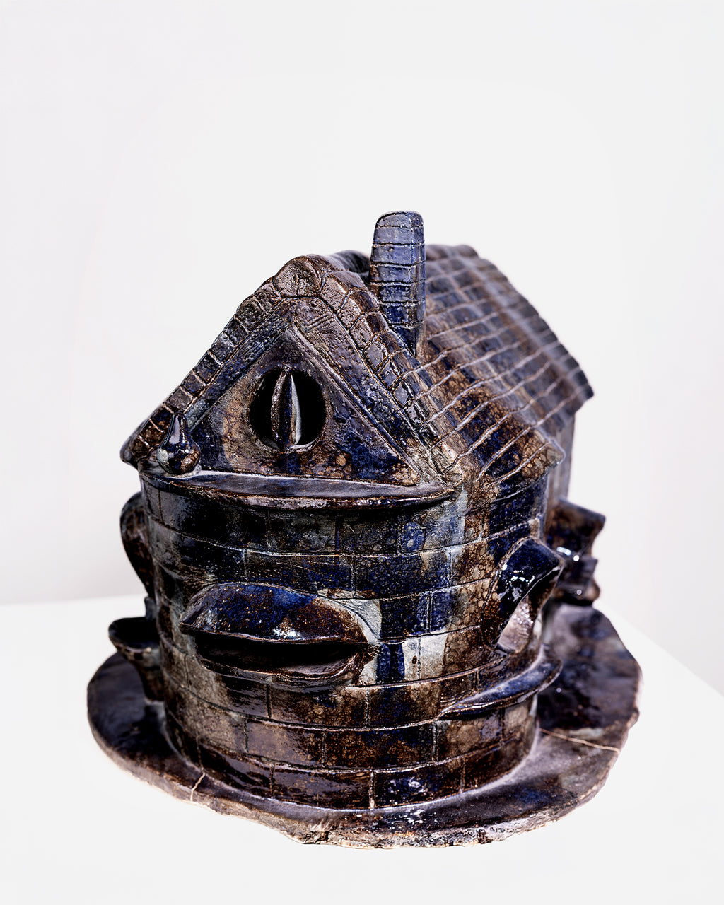 A glossy, house-shaped ceramic object with a dark blue and brown glaze. It features detailed brick-like texturing at the base, a tiled roof with a tall chimney, and a large curved barrel structure on the side, isolated against a white studio background.